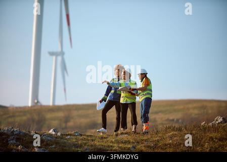 A diverse team of engineers and technicians wearing safety vests and helmets discuss plans and inspect wind turbines on a rural wind farm, collaborati Stock Photo