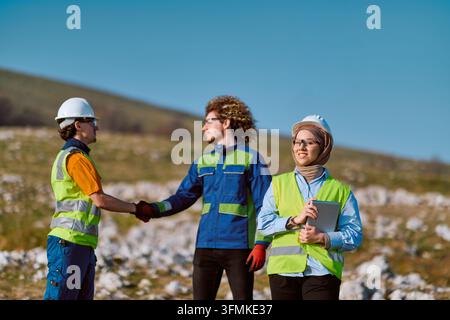 Multicultural construction team on a rural job site wearing hard hats and hi-vis vests, exchanging a handshake while a female engineer in a hijab hold Stock Photo