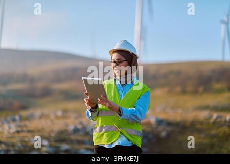A female engineer in a hard hat and high-visibility vest uses a tablet while inspecting a wind turbine site, highlighting renewable energy, field insp Stock Photo