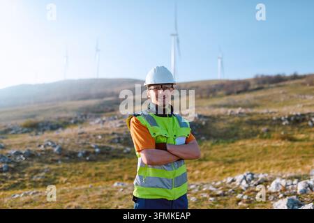 A smiling wind farm technician stands with arms crossed in a high-visibility vest and hard hat at a rural renewable energy site with wind turbines in Stock Photo
