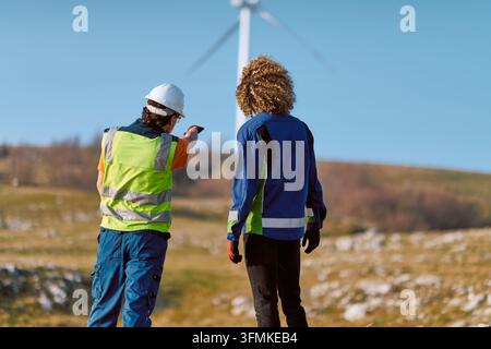 Two field technicians wearing safety vests and helmets inspect a wind turbine on a rural site, discussing maintenance, safety checks and renewable ene Stock Photo