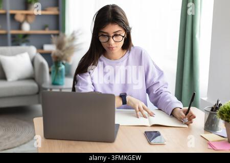 Portrait of woman in glasses taking notes while working on laptop computer at home office, free space. Freelancer noting down information from interne Stock Photo