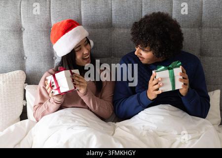 Diverse couple sitting in bed facing each other holding gift boxes, one wearing santa hat christmas Stock Photo