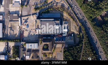 Aerial view of a large industrial facility with a flat roof, surrounding parking areas, and construction zones, situated adjacent to a multi-lane high Stock Photo
