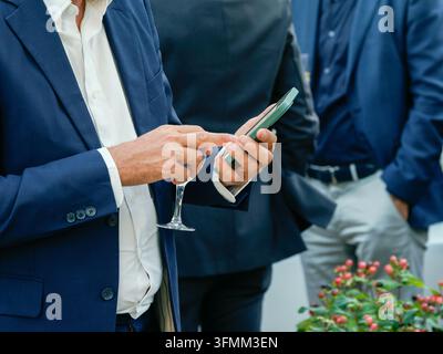 Man in blue blazer holding a glass and using a smartphone at a social event outdoors. Stock Photo