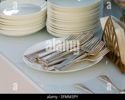 Stack of white plates and silver forks on a glass surface with a wooden napkin holder nearby. Stock Photo