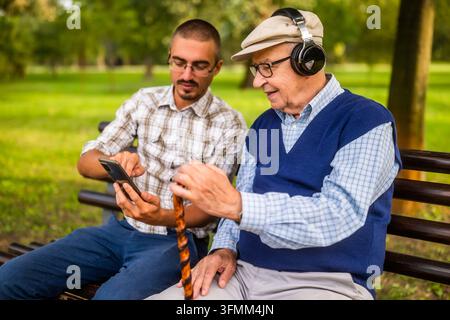 Portrait of happy grandfather and grandson in park. They are spending time together and having fun. Old man is listening music. Stock Photo