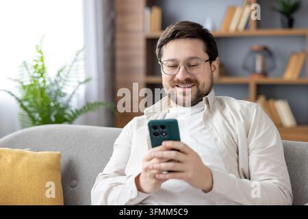 Young adult man wearing glasses sitting comfortably on a gray sofa at home, smiling while engaging with his mobile phone, communicating and interacting online Stock Photo