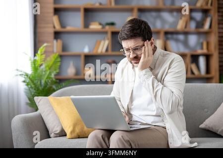 Frustrated man feeling stressed and suffering from a headache while concentrating on his laptop, working from home on a modern couch with a bookshelf in the background Stock Photo
