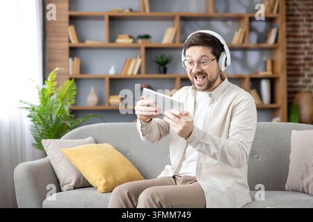 Man wearing headphones and glasses sitting on a sofa in his living room, enthusiastically playing a video game on a digital tablet, immersed in online entertainment and technology Stock Photo