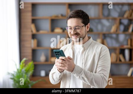 Young man wearing glasses and a casual shirt smiling while holding and interacting with a smartphone, standing indoors with a blurred bookshelf in the background Stock Photo