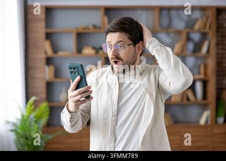 Man with glasses reacting with surprise and disbelief to unexpected information while holding a mobile phone, expressing a strong emotion of being stunned by digital content Stock Photo