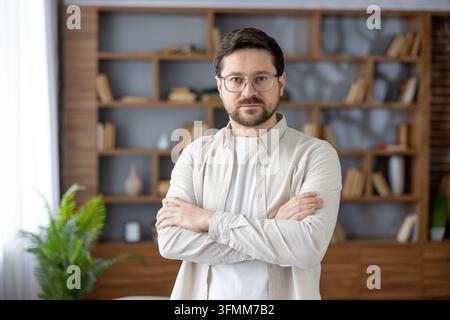 Confident bearded man with glasses stands with folded arms in a modern home office by a bookshelf, projecting expertise, leadership and reliable professional success Stock Photo