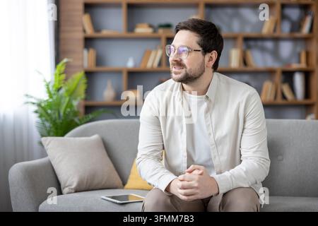Young man sitting comfortably on a gray sofa in his living room, looking away thoughtfully while planning or imagining positive outcomes, highlighting concepts of home comfort and relaxation Stock Photo