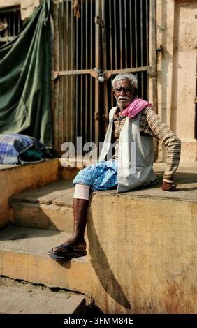 VARANASI, INDIA - JANUARY 22, 2026: An elderly man in a striped sweater, vest and checkered lungi sits on a stone ledge beside steps Stock Photo