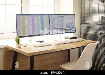 Office workspace desk with computer monitors and analytics. Bright interior by window with dual screens, charts, keyboard, mouse, plant and white chai Stock Photo