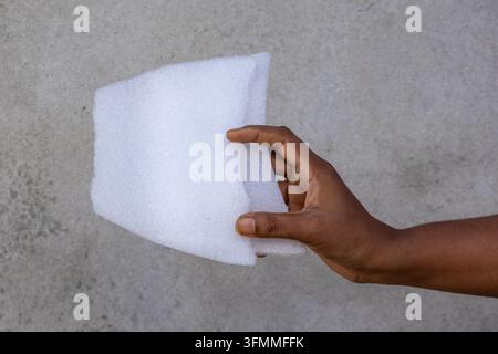 Close-up side view of a human hand holding a white poly foam sheet against gray concrete wall background. Stock Photo