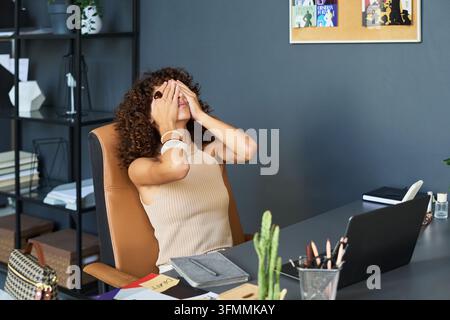 Tired Young Woman Designer Covering Eyes with Hands in Creative Office Stock Photo