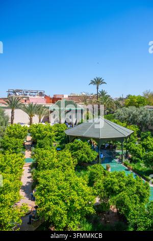 People walk and relax in a garden full of plants and trees. The sun shines brightly in the sky above. Visitors enjoy the beauty of nature in Marrakesh on a warm day. Stock Photo