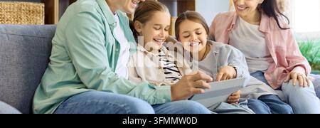 Family parents daughters watching tablet together at home. Close view of smiling faces sharing the device, browsing media and learning in a cozy room. Stock Photo