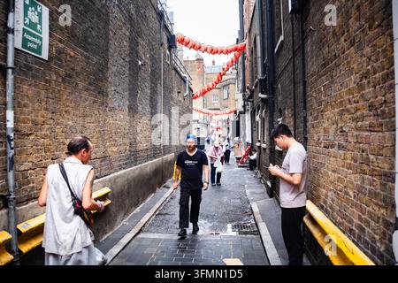 NARROW ALLEY IN LONDON'S CHINATOWN, LONDON CHINATOWN, TRAVEL REPORTAGE, LONDON, ENGLAND: People walk through a narrow brick-walled alleyway in London's Chinatown, with one man stopping to look at his phone. INFO: London’s Chinatown, nestled in the vibrant heart of Soho, is a bustling cultural hub famous for its authentic Asian restaurants, traditional bakeries, and neon-lit streets. The iconic red lanterns offer a perfect backdrop for food lovers. Stock Photo