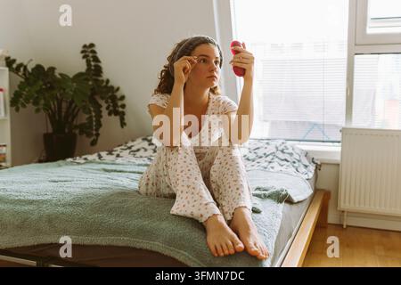 Young woman sitting on the bed in her bedroom in pyjamas, view of face, teeth, mouth in mirror Stock Photo
