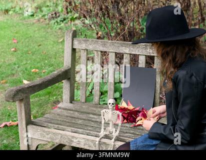 Side view of a girl wearing a black fedora and leather jacket sitting on a wooden bench, preparing a halloween display. Stock Photo