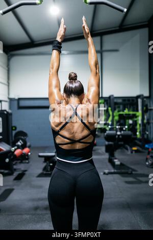 Muscular woman seen from behind performs an overhead stretch beneath a pull-up bar in a modern gym, wearing black activewear during a focused strength Stock Photo