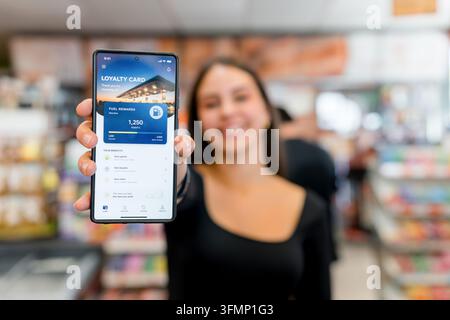 Smiling woman holds a smartphone displaying a fuel rewards loyalty card app inside a gas station convenience store, highlighting digital payment, poin Stock Photo