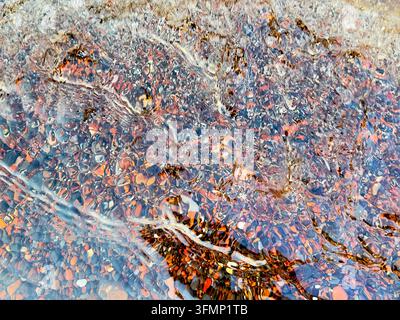 Close-up view of clear seawater flowing over colorful stones and rippled sand at a beach, creating an abstract natural texture with reflections, refra Stock Photo
