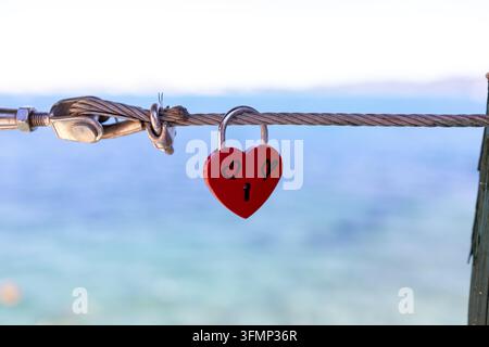 Red heart shaped love padlock hanging from a steel cable above the sea with hand drawn hearts and keyhole creating a romantic coastal symbol against s Stock Photo