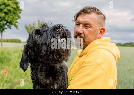 Middle-aged man in a yellow hoodie stands closely with his large black dog in a green rural field, sharing a calm moment beneath a cloudy summer sky. Stock Photo