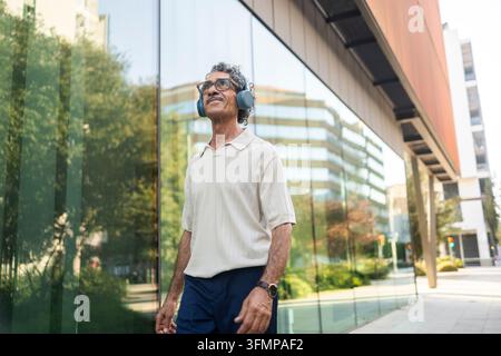 Smiling middle-aged man wearing headphones and casual clothing walks beside reflective glass offices in a modern city, enjoying music during an urban Stock Photo