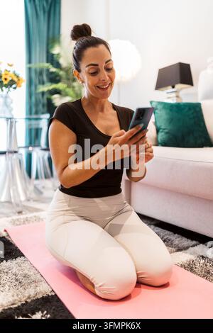 Smiling woman kneels on a yoga mat in her living room, using a smartphone after home exercise. She wears activewear beside a sofa in a bright modern i Stock Photo
