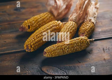 Close-up of dried corn cobs with golden grain kernels arranged on a dark rustic wooden table, creating a warm agricultural harvest and natural food ba Stock Photo