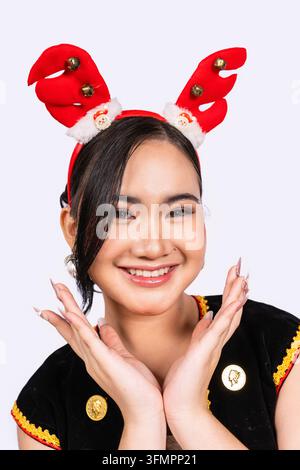An extreme close up portrait of a smiling young Sabahan woman framing her face with both hands while wearing red Christmas reindeer antlers and tradit Stock Photo