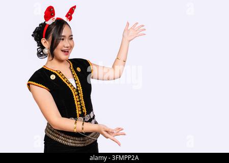 A happy young Sabahan woman in traditional Kadazan Dusun dress looks and presents toward copy space on the right while wearing red Christmas reindeer Stock Photo