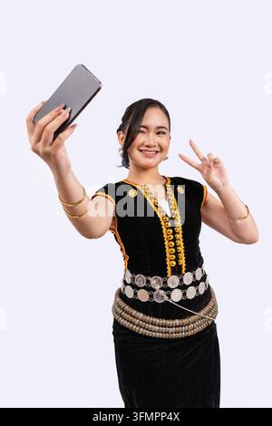 A playful young Sabahan woman in traditional Kadazan Dusun dress winks and shows a peace sign while taking a selfie with her smartphone. Stock Photo