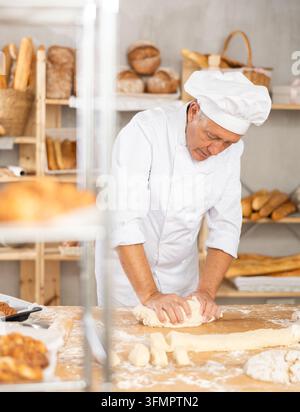 Senior man works in bakery as baker, kneads dough, works with flour. Working moment, process of creating croissants in bakery Stock Photo
