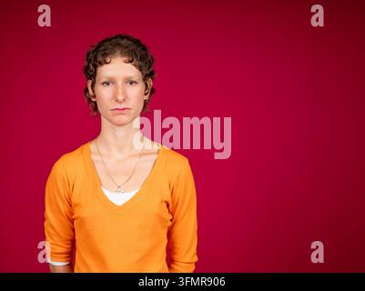 Serious young woman with short curly hair and eyebrow piercing posing against a vibrant red background, copy space Stock Photo