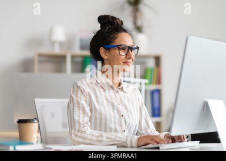 Smiling Hispanic Female Working At Computer In Modern Office, Business Concept Stock Photo