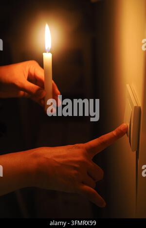 A man holds a candle while reaching for a light switch in a dark room. electricity crisis concept. Stock Photo