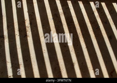 High-angle view of strong, parallel lines of light and shadow cast across a dark ground surface, creating an abstract geometric pattern Stock Photo