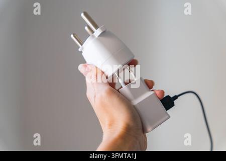 Hand holds a white travel plug adapter with three round Type M pins connected to a black charging cable. Electrical accessory for powering devices Stock Photo