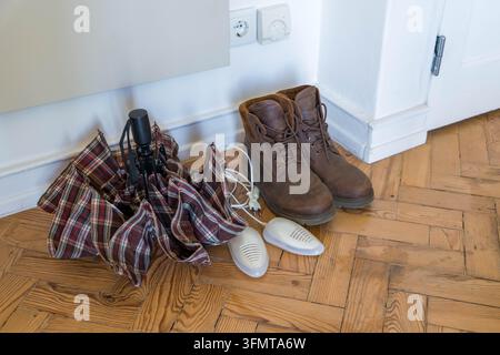 Electric shoe dryers and wet leather boots on a parquet floor beside a folded umbrella in a hallway. Autumn footwear care and drying of damp shoes Stock Photo