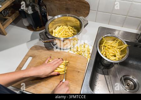 Hands trim fresh yellow beans on a wooden cutting board beside a pot and colander in a home kitchen, preparing vegetables for a homemade dinner meal. Stock Photo