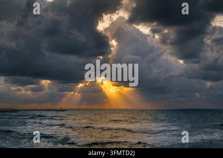 Dramatic seascape with dark storm clouds gathering above the ocean as golden sunlight breaks through openings in the sky. Powerful sun rays illuminate Stock Photo