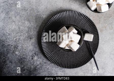 Overhead view of soft white marshmallows in a black bowl on a ribbed black plate with spoon, set against a textured gray stone background, with extra Stock Photo