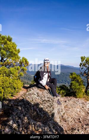 Smiling woman hiker in a helmet and sunglasses rests on a rocky mountain summit trail beside pine trees, with forested ridges and a clear blue sky in Stock Photo