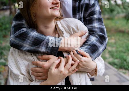 Close-up of a smiling young woman embraced from behind by her partner in a green park, expressing romantic affection, trust, tenderness, and emotional Stock Photo